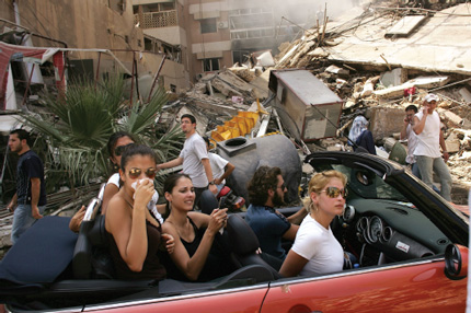 4 women and one man sit in a parked car, the look out of the car at the devistation from bombing around them.