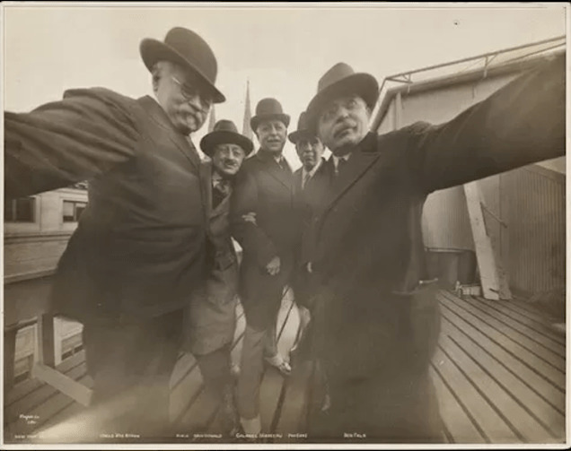 Fish eye distorted selfie of 5 suited men in hats. Image is off yellow monochrome and appears to have been taken in the 1930s.